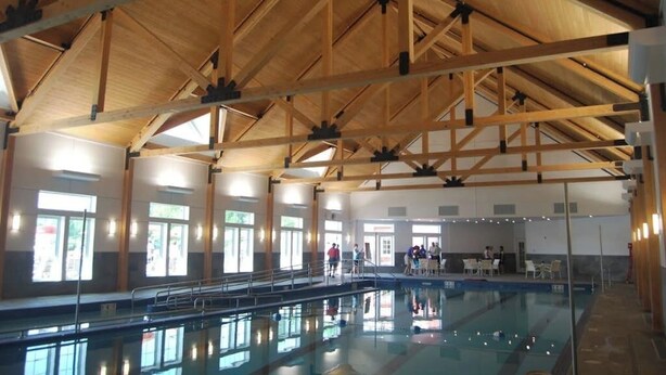 Una piscina interior con vigas de madera, ventanales amplios y suelo de baldosas.
