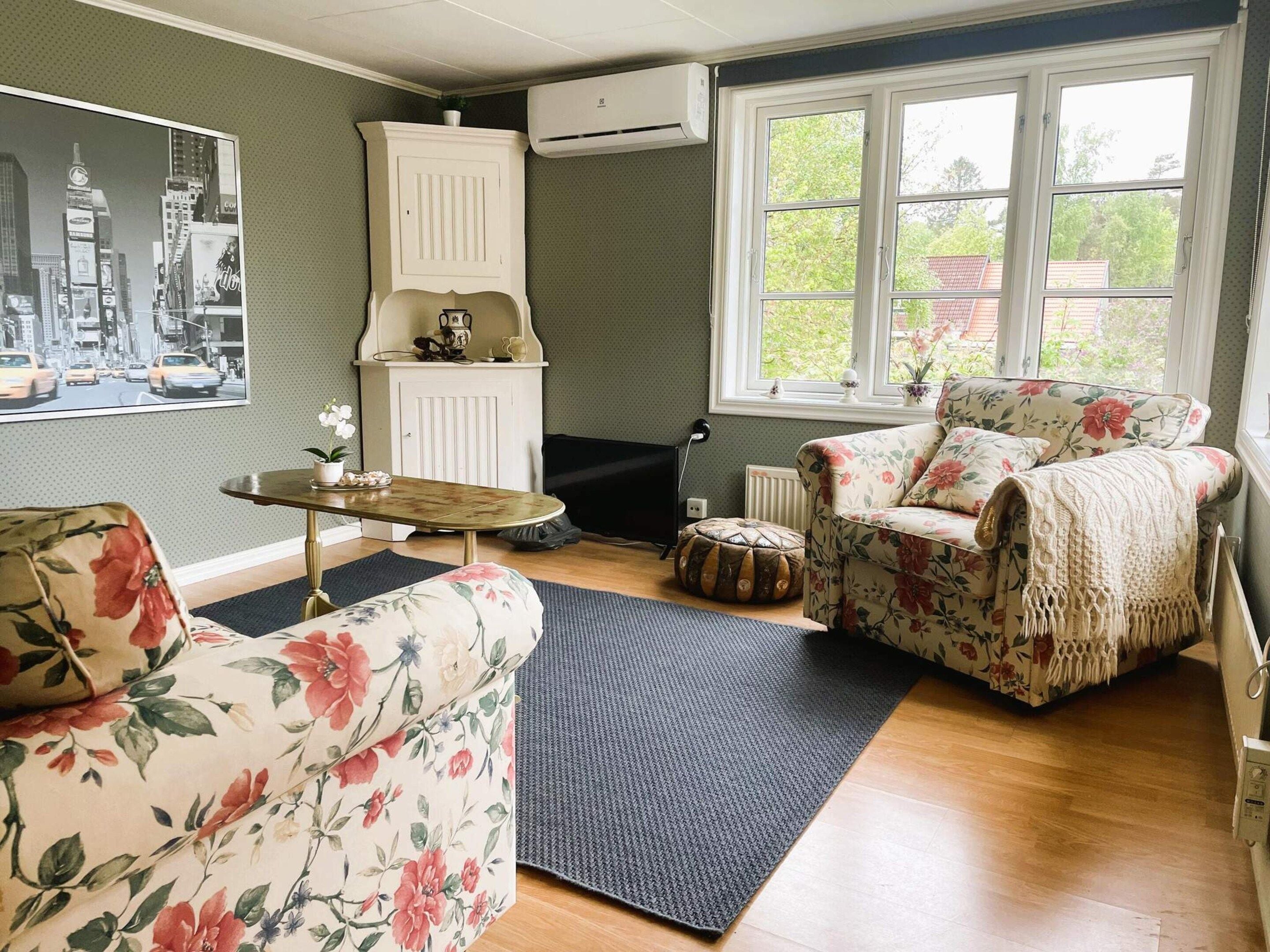 Een woonkamer met bloemenmotieven op de bekleding, een ronde salontafel, een bank, een kast en een groot raam met uitzicht op groen.