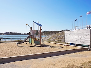 Une aire de jeux de plage avec un toboggan et une structure d’escalade, une clôture en bois et des drapeaux.