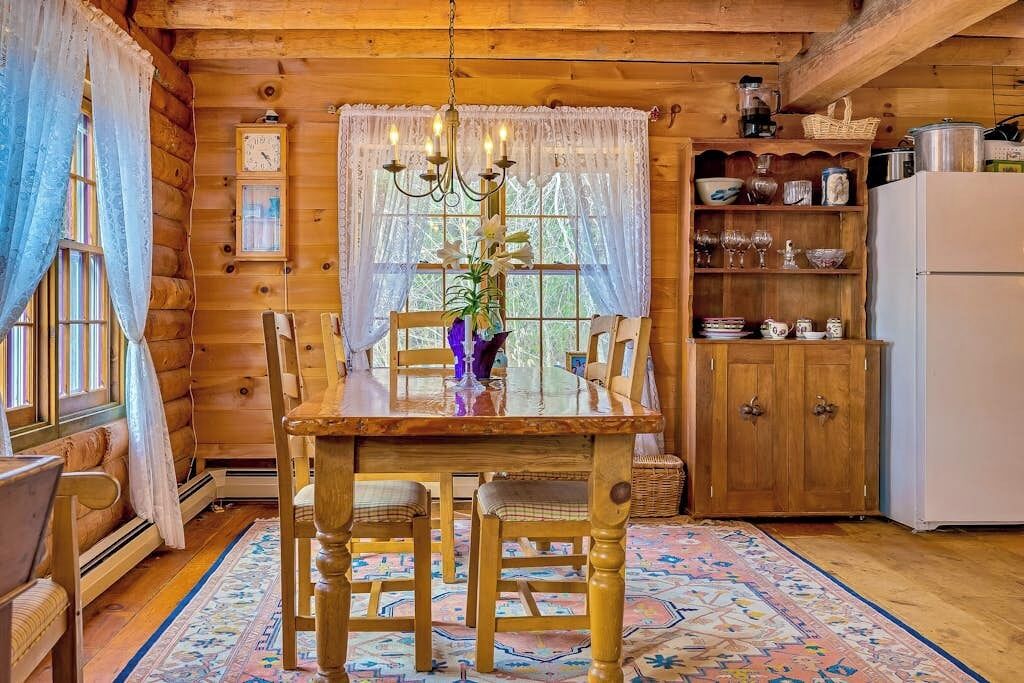 L'intérieur d'un chalet en bois, avec une table à manger, des chaises, un lustre, une horloge et un buffet contenant de la vaisselle.