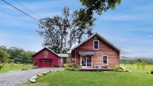 A two-story wooden house with a red garage, a porch with chairs, and a grassy yard. - Historic home on 1 acre with snowmobile trails in backyard & mountain views (Waitsfield)