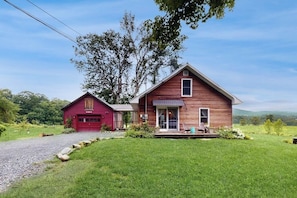 Une maison en bois à deux étages, avec un garage rouge, une véranda avec des chaises et une cour couverte d’herbe.