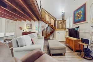A living room with a sofa, ottoman, wooden staircase, TV, and artwork on the wall. - Walk-to-village end-unit condo, mountain views, large deck, woodstove, bbq, a/c (Quechee)