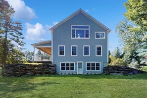 A two-story house with a stone foundation, a porch, and a clear sky. - Private house w/ seasonal mountain & meadow views, pond, balcony, porch, jacuzzi (Waitsfield)