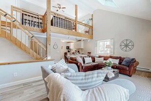 A spacious living area with a high wooden staircase, a cozy seating area with a brown leather sofa, and a kitchen in the background.