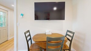 Un coin repas avec une table en bois et quatre chaises, un téléviseur à écran plat fixé au mur, et une porte donnant sur une fenêtre.