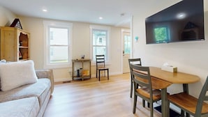 A living room with a sofa, a dining table with chairs, a TV mounted on the wall, and a window. - Centrally located apartment for the skier & outdoor enthusiast (Rutland)