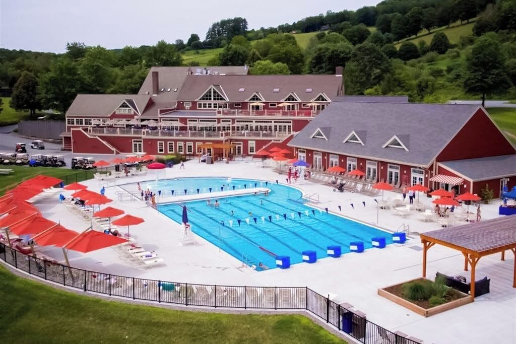 Una amplia piscina con múltiples carriles, rodeada de sillones de descanso y sombrillas, con un edificio de fachada roja y una zona cubierta al frente.