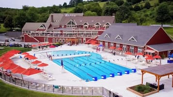 A large swimming pool with multiple lanes, surrounded by lounge chairs and umbrellas, with a building featuring a red facade and a covered area in the foreground.