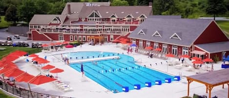 Una amplia piscina con múltiples carriles, rodeada de sillones de descanso y sombrillas, con un edificio de fachada roja y una zona cubierta al frente.