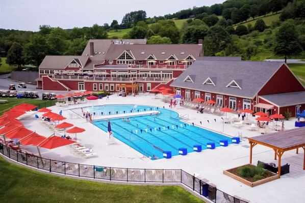 A large swimming pool with multiple lanes, surrounded by lounge chairs and umbrellas, with a building featuring a red facade and a covered area in the foreground.