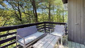 Une terrasse en bois avec deux bancs blancs et une petite table, entourée d’arbres.