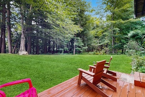 A wooden deck with a red chair and a pink chair, surrounded by a well-maintained lawn and dense trees. - End-unit condo w/ fireplace, pool, sauna, tennis, deck, ski locker, w/d (Waitsfield)