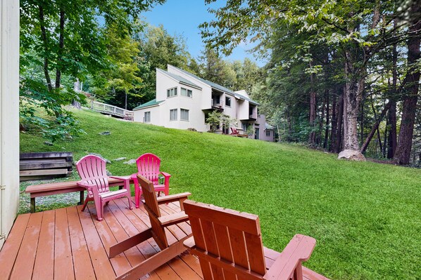 A wooden deck with Adirondack chairs and a bench, overlooking a grassy hill with a white building.