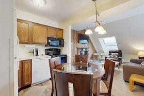 A compact kitchen with wooden cabinets, a microwave, and a stove. There's a dining area with a glass table and chairs. A living room with a sofa and a TV is visible in the background. - Top-Floor Condo w/ view of Killington ski slopes, shared pool, hot tub, sauna (Killington)