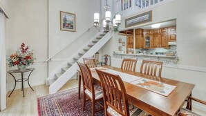 Un área de comedor con una mesa de madera, sillas, un tapete, una escalera y una cocina al fondo.
