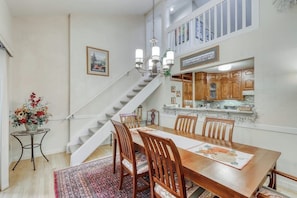 Un área de comedor con una mesa de madera, sillas, un tapete, una escalera y una cocina al fondo.