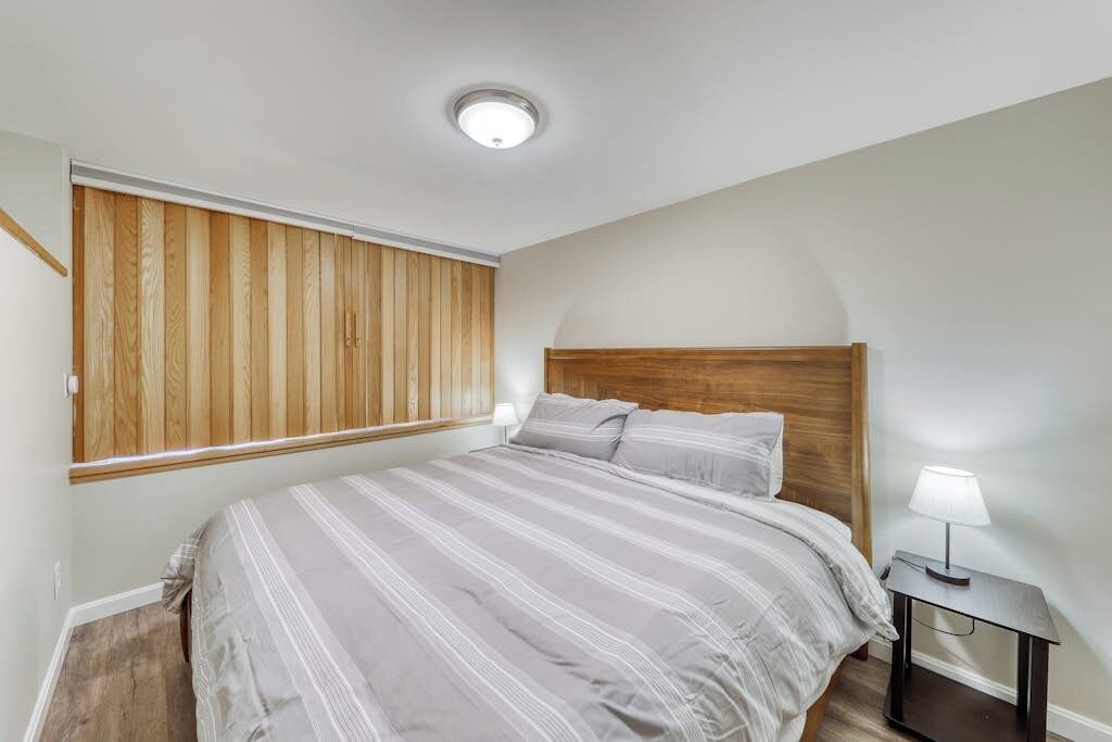 Un dormitorio con cama, mesita de noche, lámpara, cabecera de madera y ventana con persianas.