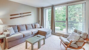 A living room with a sofa, a coffee table, a chair, and a large window with a view of greenery. - Ski-In/Ski-Out Second-Floor Condo w/ Free WiFi, Mountain Views, & a Shared Pool (Warren)