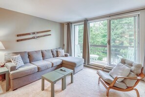 A living room with a sofa, a coffee table, a chair, and a large window with a view of greenery. - Ski-In/Ski-Out Second-Floor Condo w/ Free WiFi, Mountain Views, & a Shared Pool (Warren)