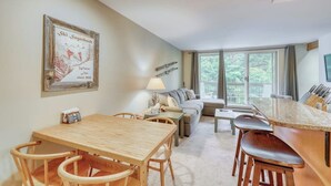 A dining area with a wooden table and chairs, a sofa, and a large window with a view of greenery. - Ski-In/Ski-Out Second-Floor Condo w/ Free WiFi, Mountain Views, & a Shared Pool (Warren)