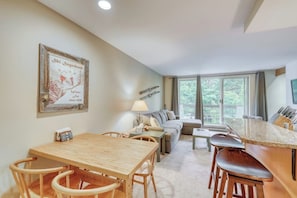 A dining area with a wooden table and chairs, a sofa, and a large window with a view of greenery. - Ski-In/Ski-Out Second-Floor Condo w/ Free WiFi, Mountain Views, & a Shared Pool (Warren)