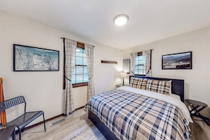 Ein Schlafzimmer mit einem Bett, zwei Vorhängen an den Fenstern, einem Sessel, einem kleinen Tisch und zwei gerahmten Bildern an der Wand.