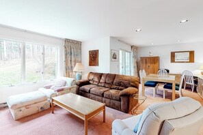 A spacious living room with a brown leather sofa, a white armchair, a wooden coffee table, and a dining area with a wooden table and chairs. - Ski condo with locker, shuttle, fast WiFi, fireplace, washer/dryer & AC (Warren)