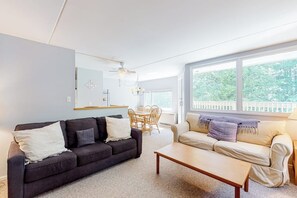 Ein Wohnzimmer mit einem dunkelgrauen Sofa, einem hellen Sofa, einem Holztisch und einer Essecke mit Tisch und Stühlen. Ein großes Fenster bietet Blick ins Grüne.