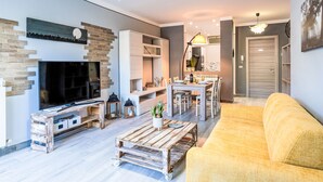 Een moderne woonkamer met een flatscreen televisie, een houten salontafel, een gele bank en een eethoek met tafel en stoelen.