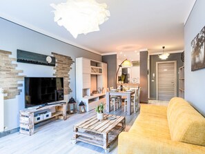 Ein modernes Wohnzimmer mit Flachbildfernseher, Couchtisch aus Holz, gelbes Sofa und einem Essbereich mit Tisch und Stühlen.