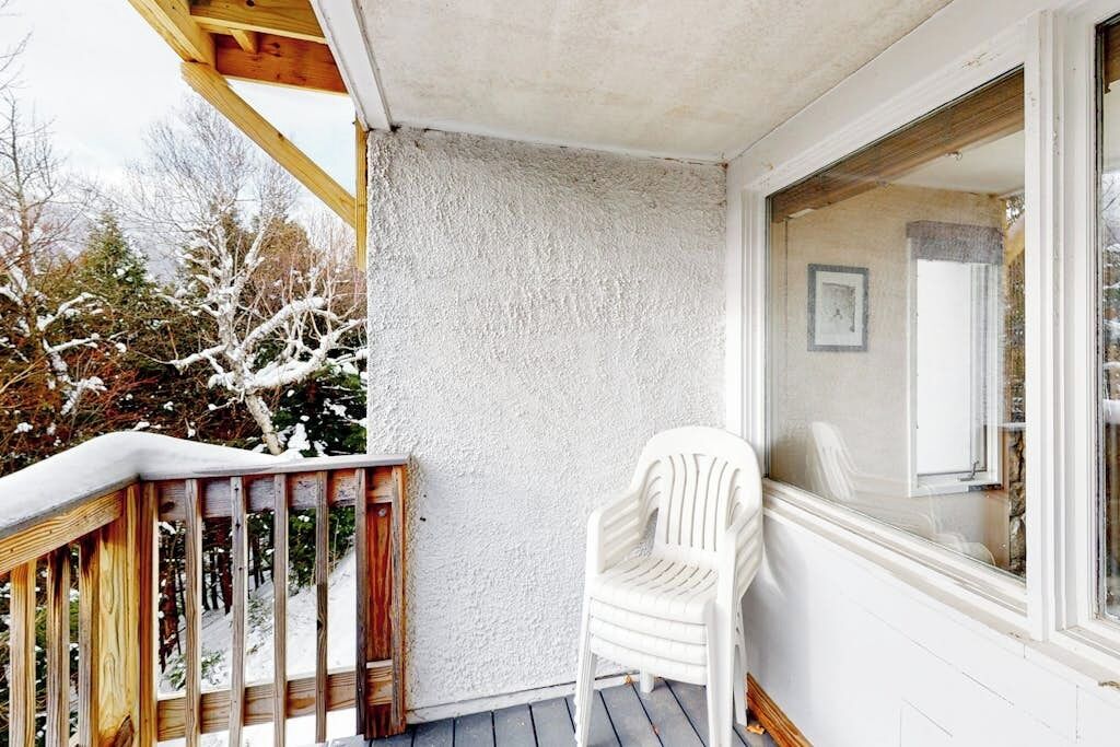 A wooden deck with a white plastic chair, a white textured wall, and a window with a framed picture.