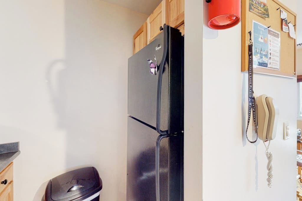 A kitchen with a black refrigerator, wooden cabinets, a trash bin, a corkboard with notes, and a telephone.