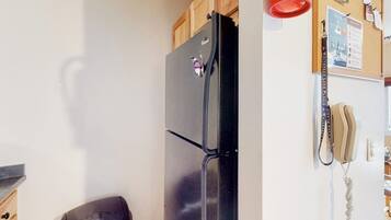 A kitchen with a black refrigerator, wooden cabinets, a trash bin, a corkboard with notes, and a telephone.