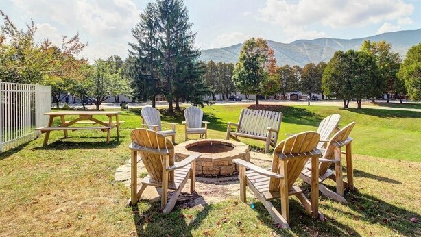 Et udendørs opholdsområde med Adirondack-stole og et bålsted, omgivet af græsplæne og træer.