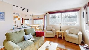 En opholdsstue med sofa, to lænestole, sofabord og et køkkenområde med bord og stole.