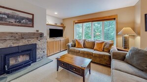 A living room with a stone fireplace, a sofa, a coffee table, a television, and a window with a view of trees.