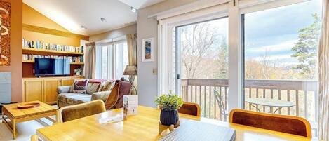 A spacious living area with a wooden dining table, comfortable seating, a bookshelf, a television, and a balcony with a view.