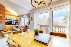 Ein geräumiger Wohnbereich mit einem Holzespeisetisch, bequemen Sitzmöglichkeiten, einem Bücherregal, einem Fernseher und einem Balkon mit Aussicht.