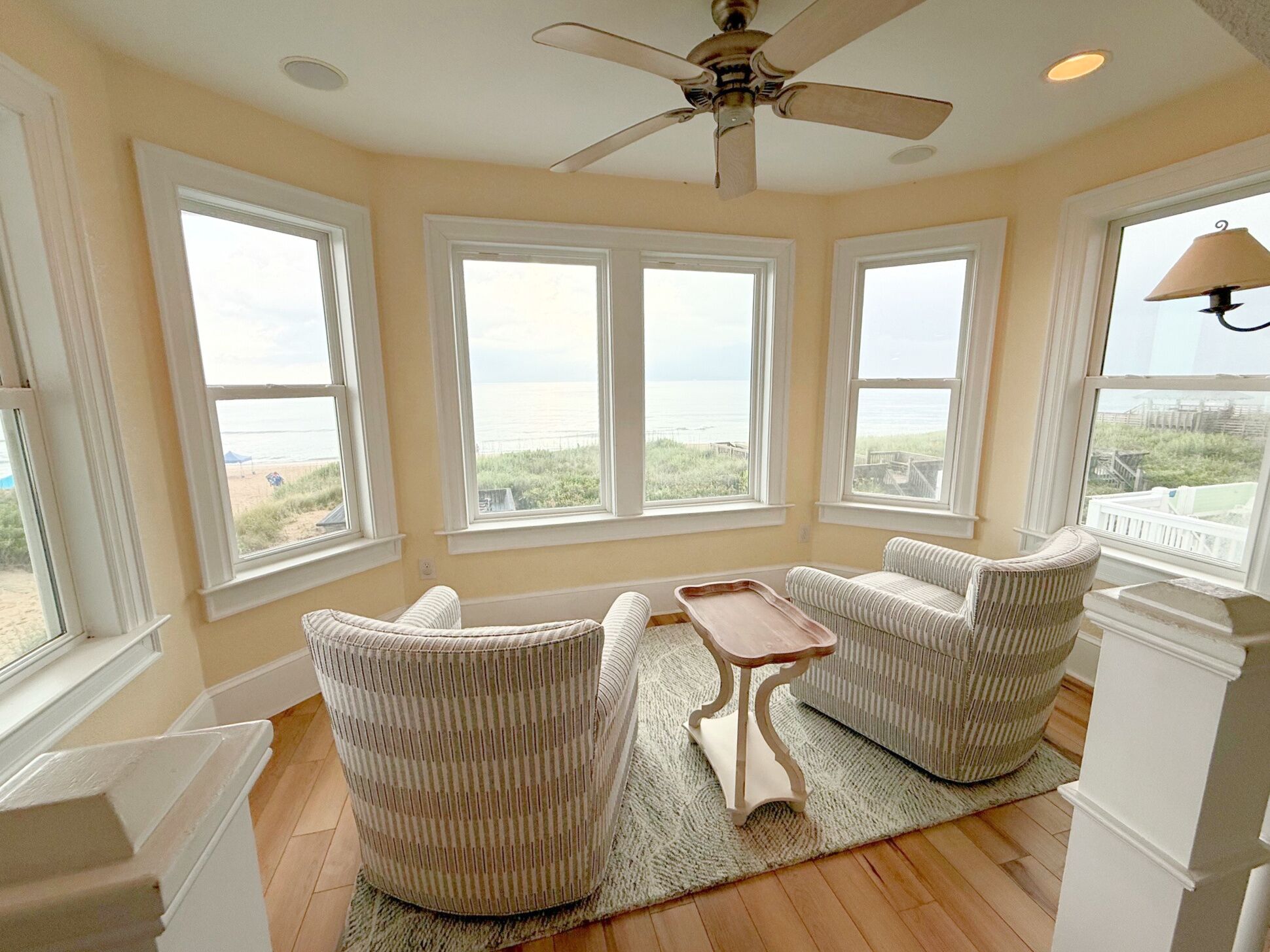 A room with large windows, a ceiling fan, and two upholstered chairs with a small table.