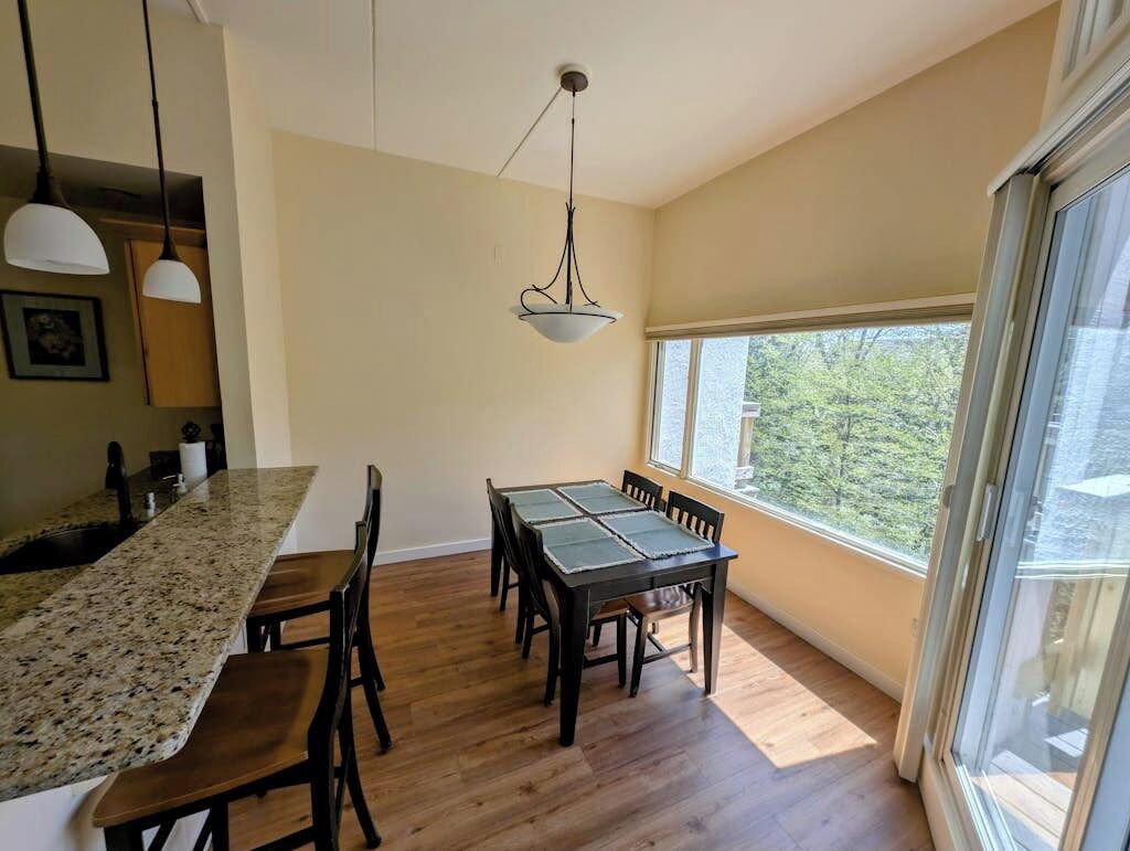A dining area with a table and chairs, a kitchen counter, pendant lighting, and a large window with a view.