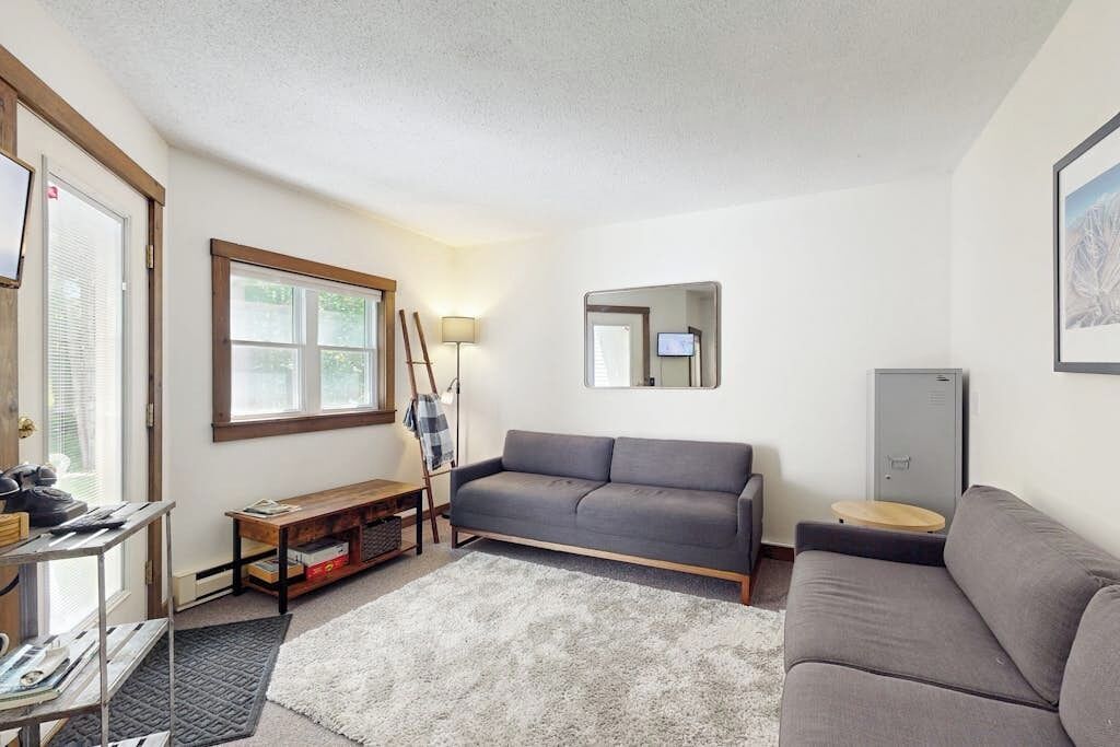 A living room with a grey sofa, a wooden bench, a small table, a lamp, and a mirror on the wall.