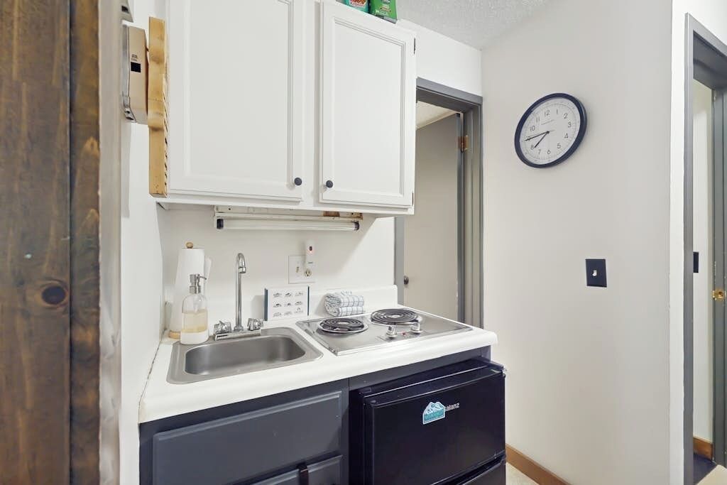 Une cuisine de taille réduite, avec des meubles blancs, un évier, une cuisinière et une horloge au mur.