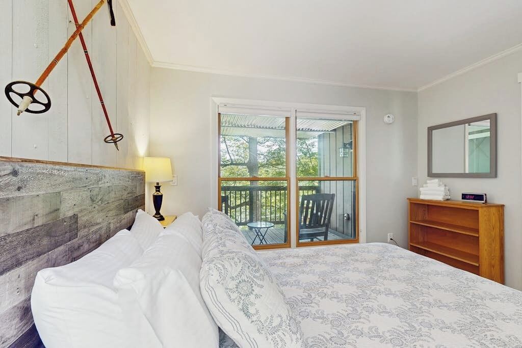 A bedroom with a bed, a wooden headboard, a nightstand with a lamp, a wooden dresser, a sliding glass door to a balcony, and a mirror on the wall.