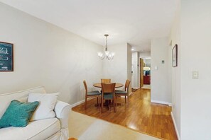 A dining area with a wooden table and chairs, a chandelier, a white sofa with a teal pillow, and a hallway leading to a kitchen. - Top floor condo w/ balcony, gas fireplace, tennis, walking/biking trails, w/d (Warren)