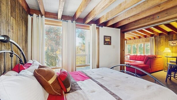 A bedroom with a bed, a window, and a view of a red couch and a lamp.