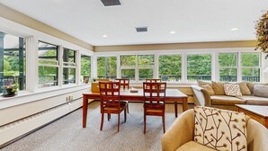 Una habitación iluminada por el sol con una mesa de comedor, sillas, un sofá y ventanas amplias que dan a vistas de vegetación.
