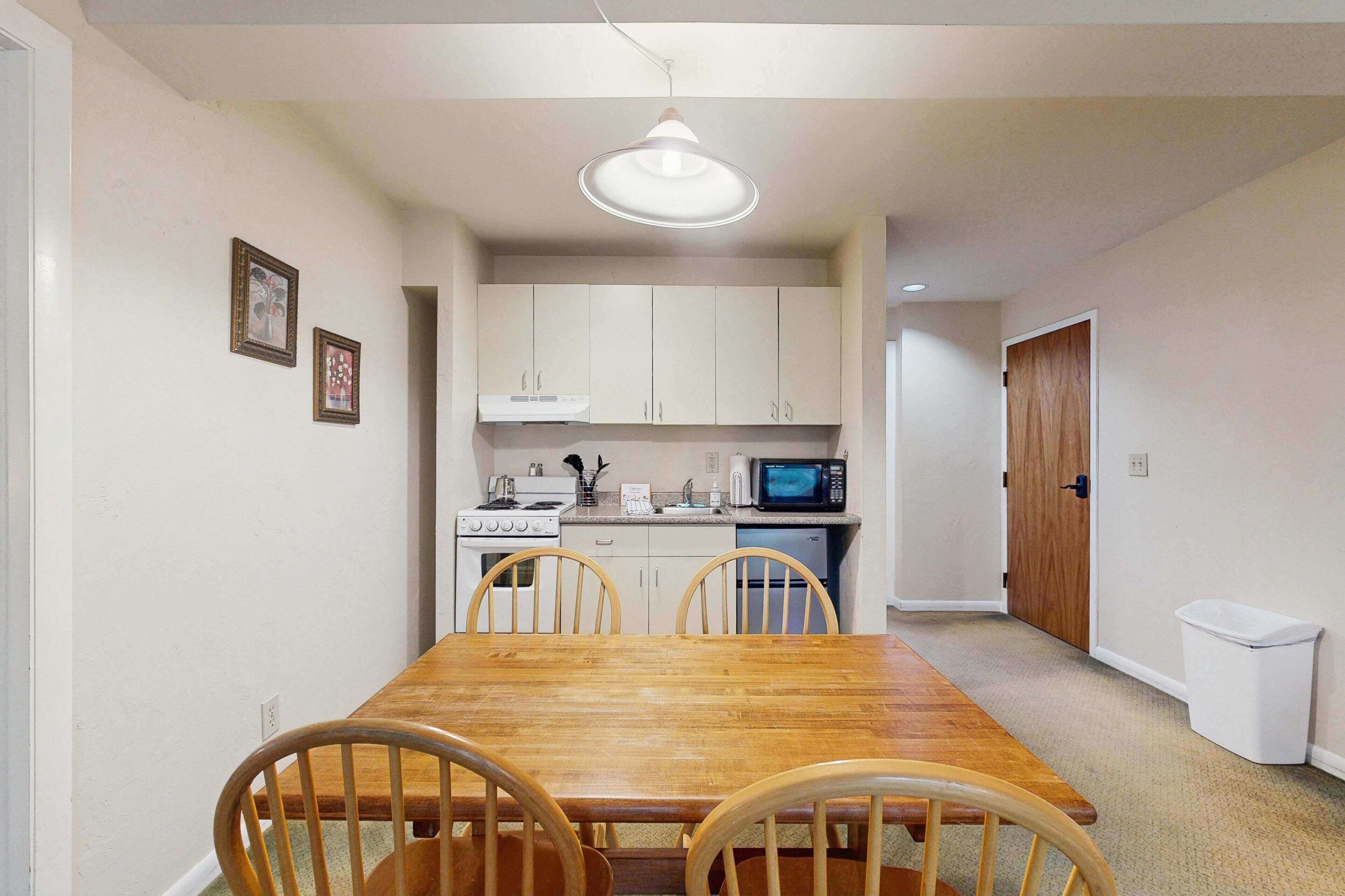 Un comedor con mesa y sillas de madera, una cocina con muebles y electrodomésticos blancos, y un cubo de basura blanco.
