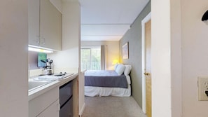 A narrow room with a bed, a desk with a lamp, and a window with curtains.