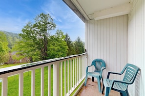 A balcony with a white railing, two blue chairs, and a view of a green landscape with trees and a house.